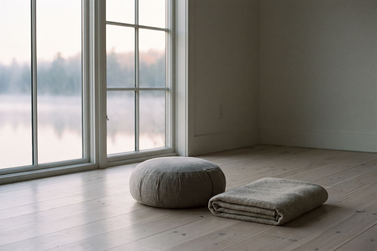 A linen meditation cushion and folded wool blanket on pale floorboards, a misty lake through a tall window.