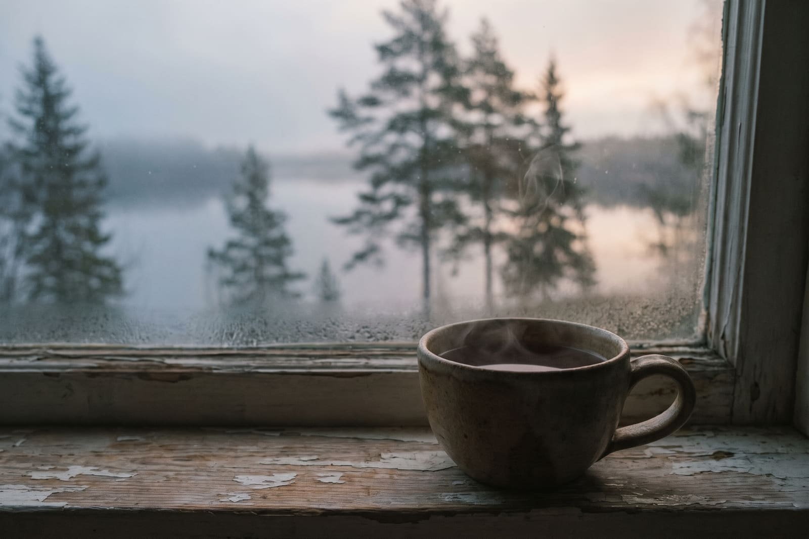 A ceramic cup of tea steaming on a weathered windowsill, a misty lake beyond the glass.